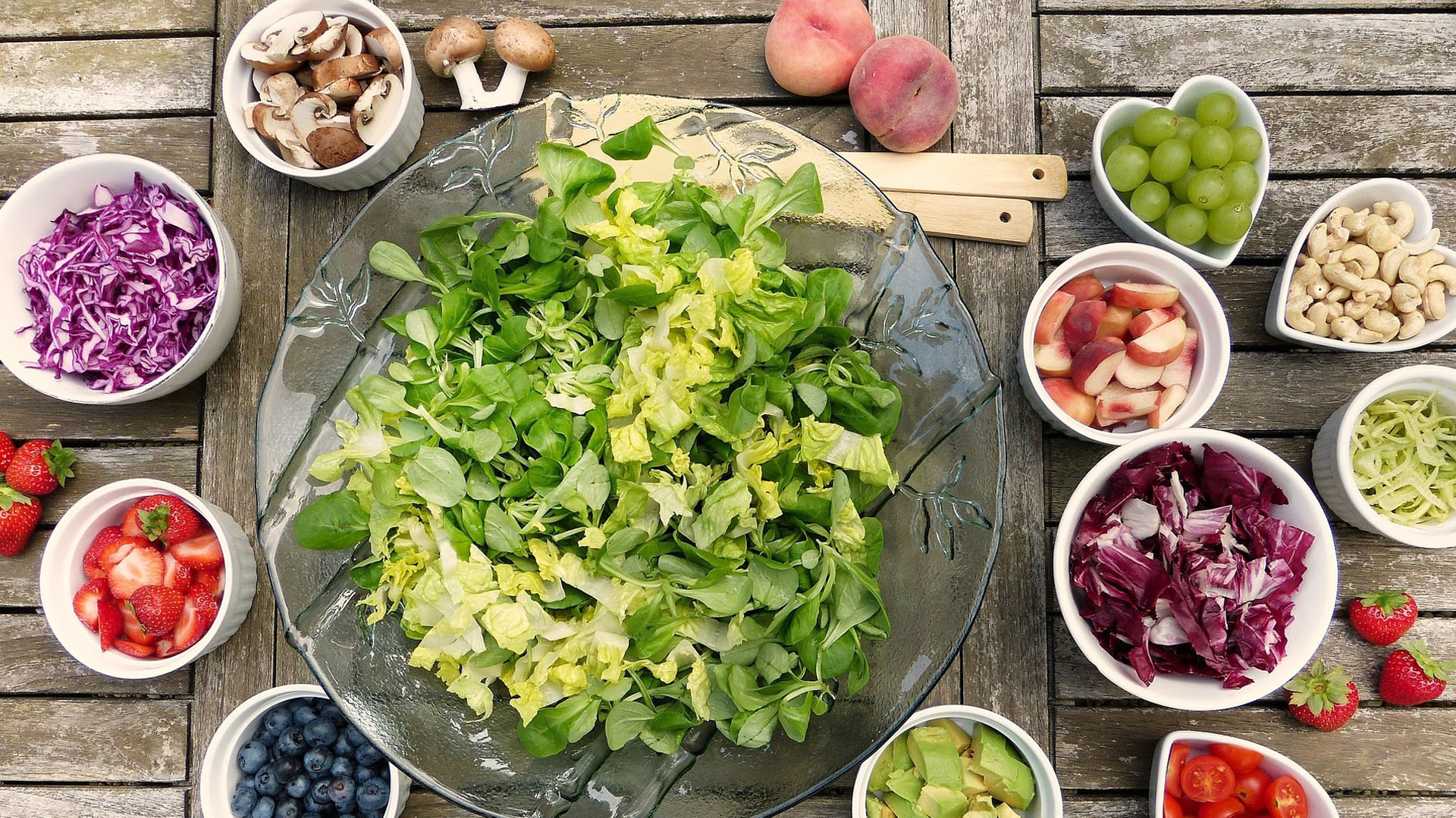 Eine große Salatschüssel in der Mitte und viele kleine Schüsseln rundherum, gefüllt mit frischem Obst, Gemüse und Nüssen.